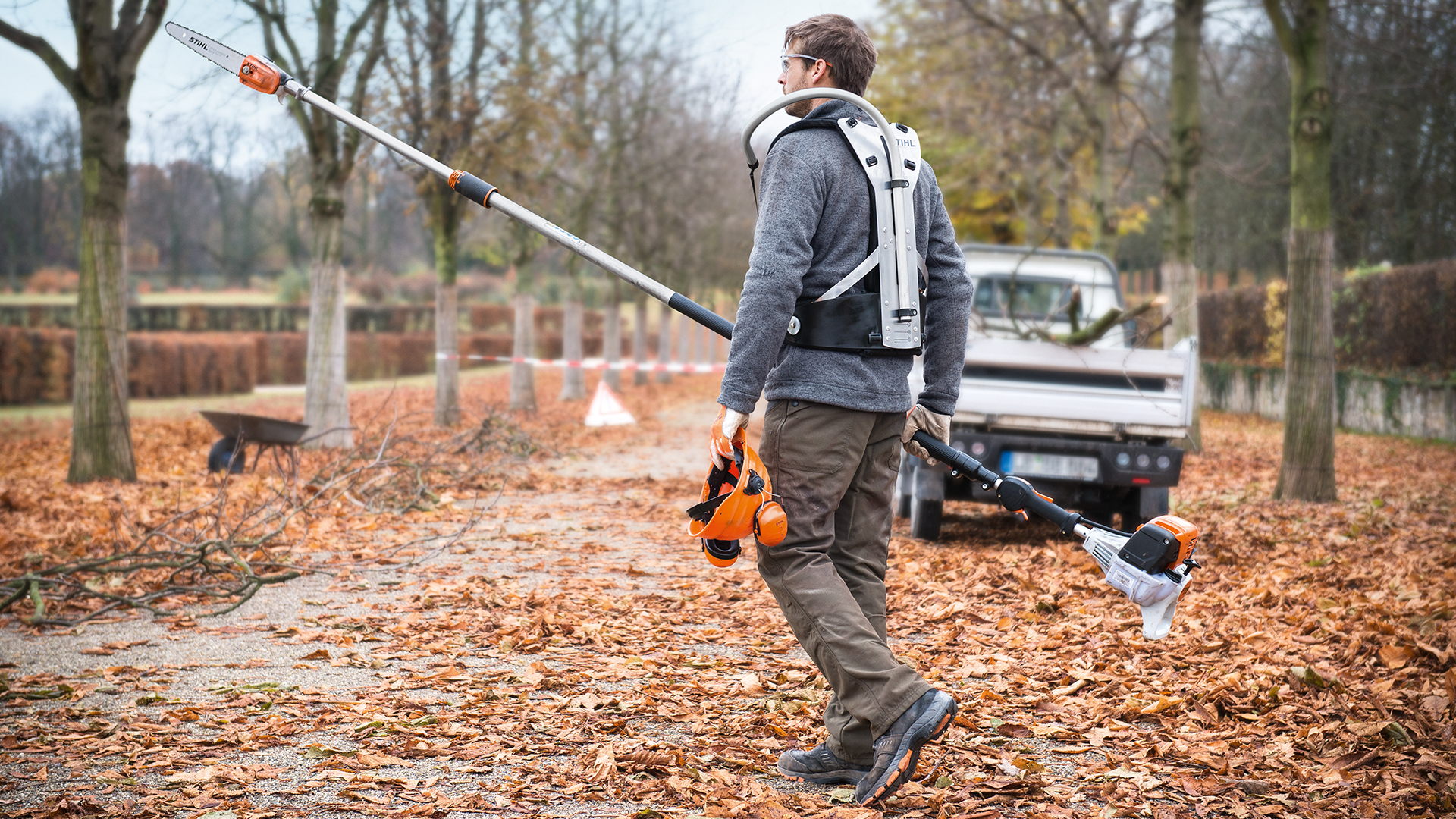 Homme équipé d'une perche élagueuse thermique STIHL HT 133 et véhicule de travail dans le parc en automne