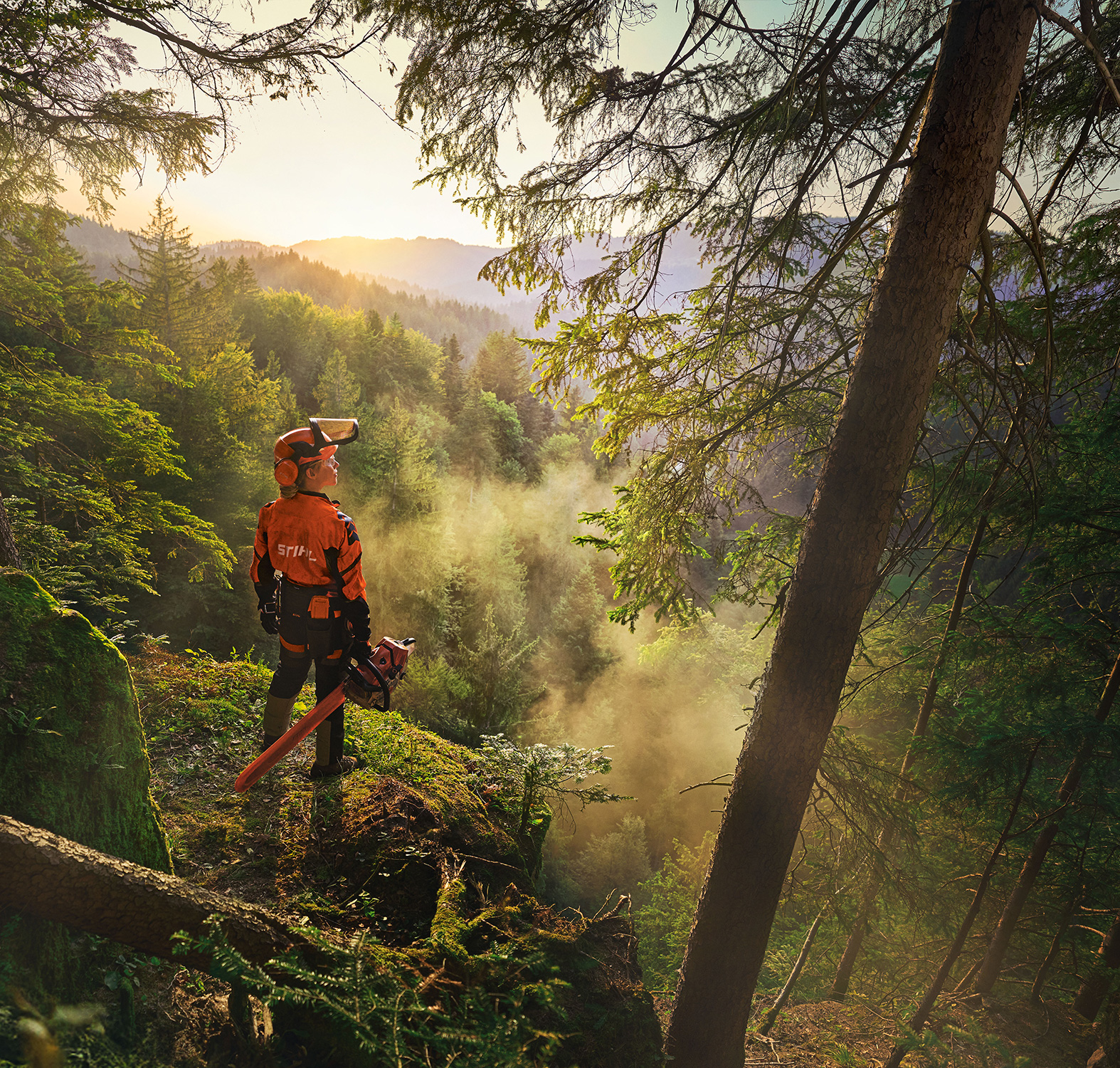 Une femme se tient au bord d'une falaise dans une forêt au vert saturé. Elle porte des vêtements de travail STIHL orange, le casque ADVANCE ProCom, et tient une tronçonneuse STIHL MS.