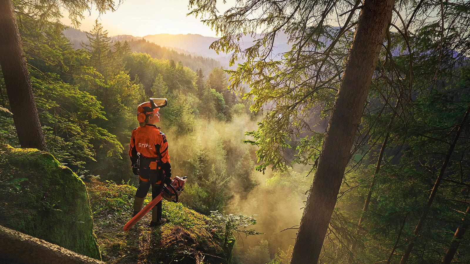 Une femme se tient au bord d'une falaise dans une forêt d'un vert saturé. Elle porte des vêtements de travail STIHL orange, le casque ADVANCE ProComHeadset, et tient une tronçonneuse STIHL MS.