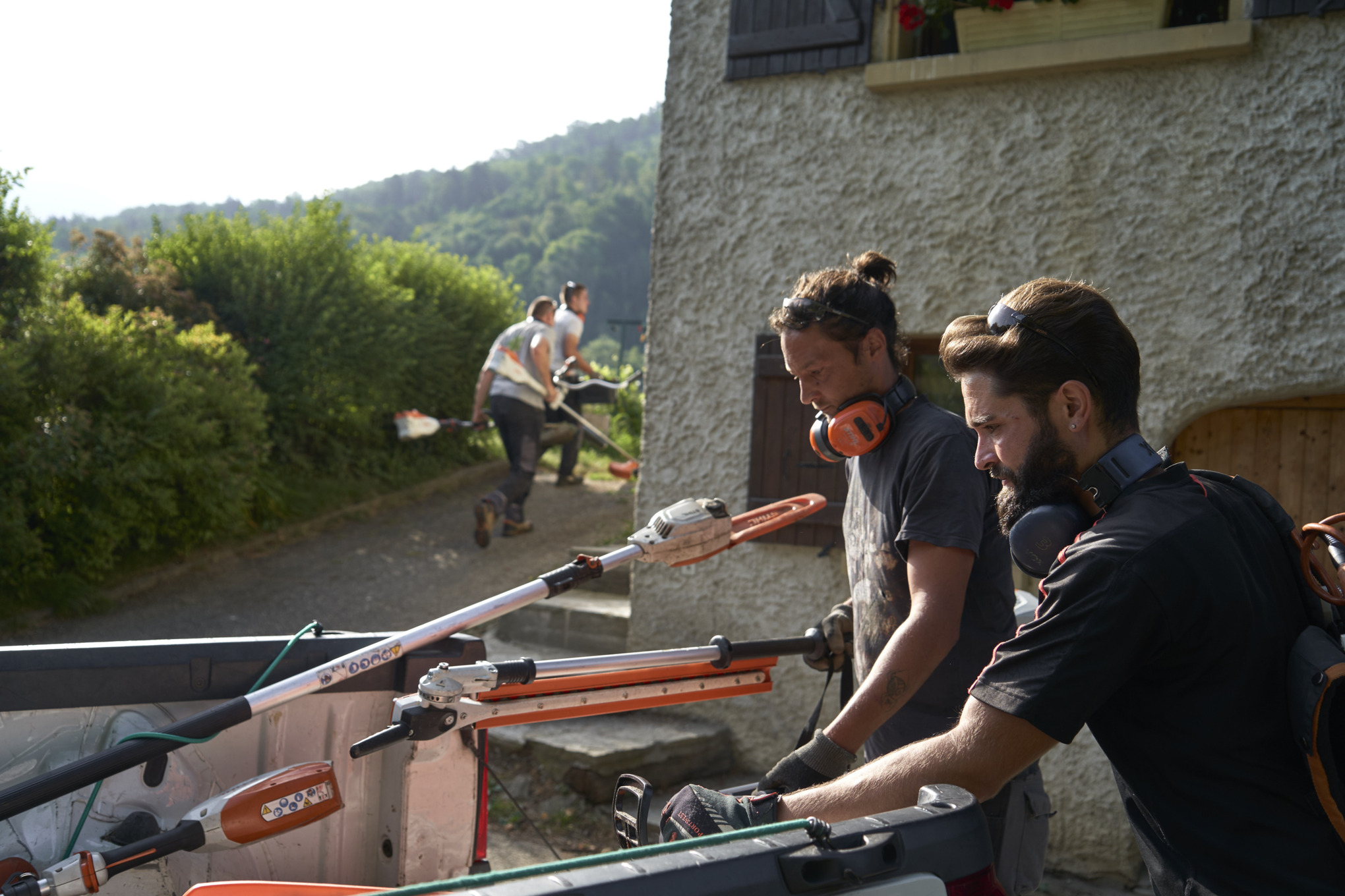 Deux paysagistes déchargeant des appareils à batterie STIHL d’une voiture et collègues en arrière-plan 