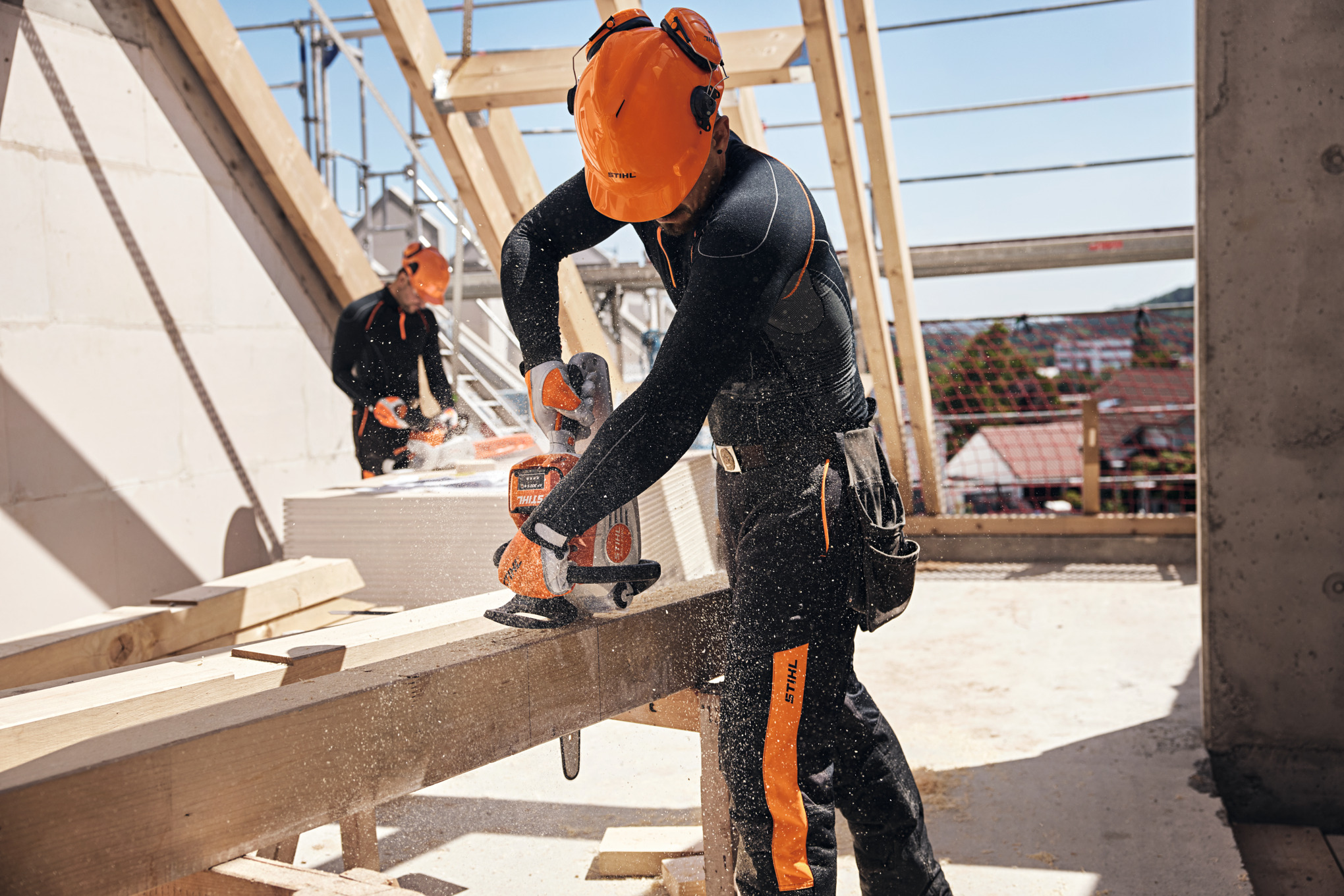 Professionnel STIHL dans une charpente de toit découpant une poutre en bois avec une tronçonneuse à batterie, aux côtés d’un autre collègue en arrière-plan, en utilisant une lui aussi