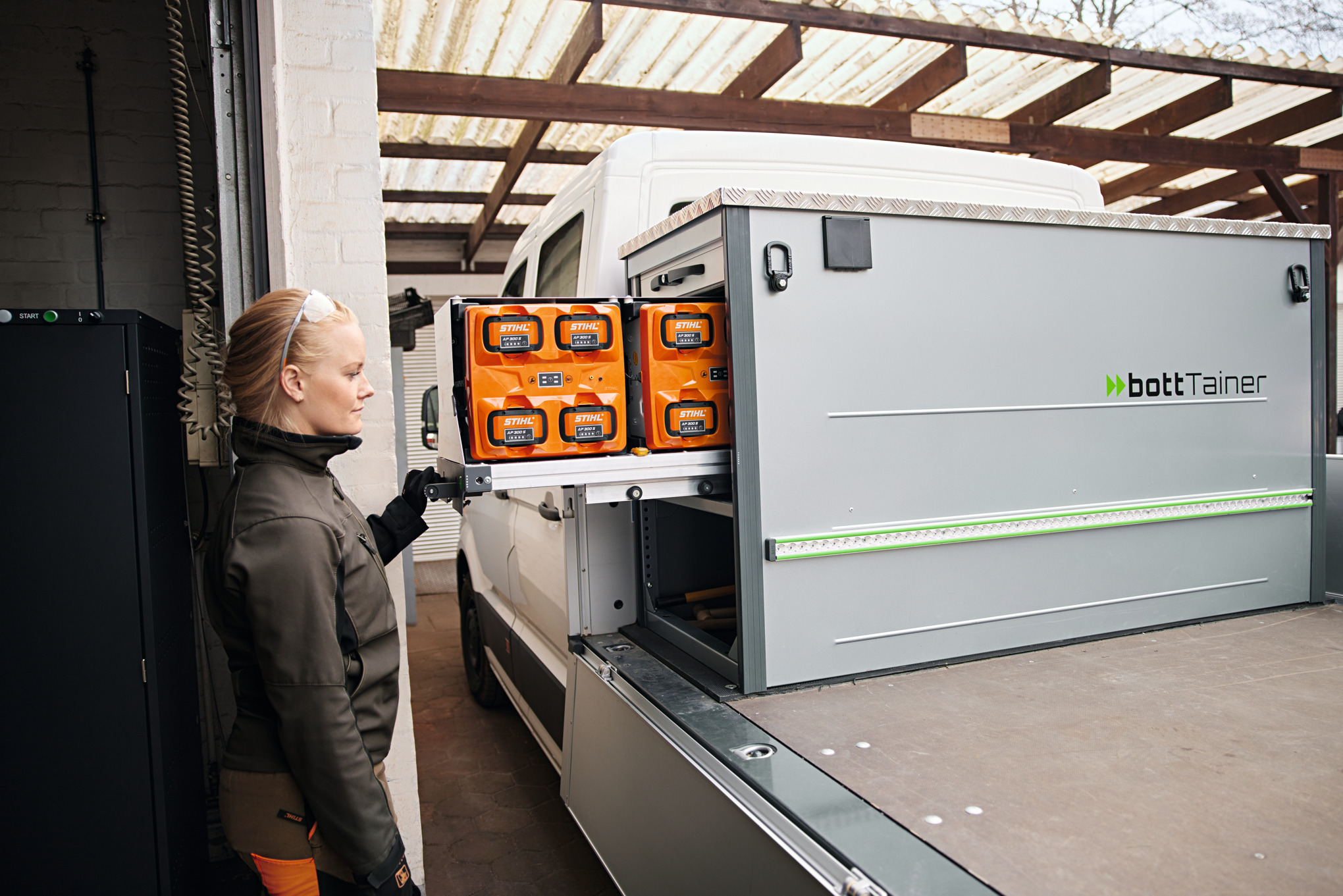 Femme blonde de profil à côté d’un véhicule avec plateau de chargement en train de manipuler les batteries dans le bottTainer