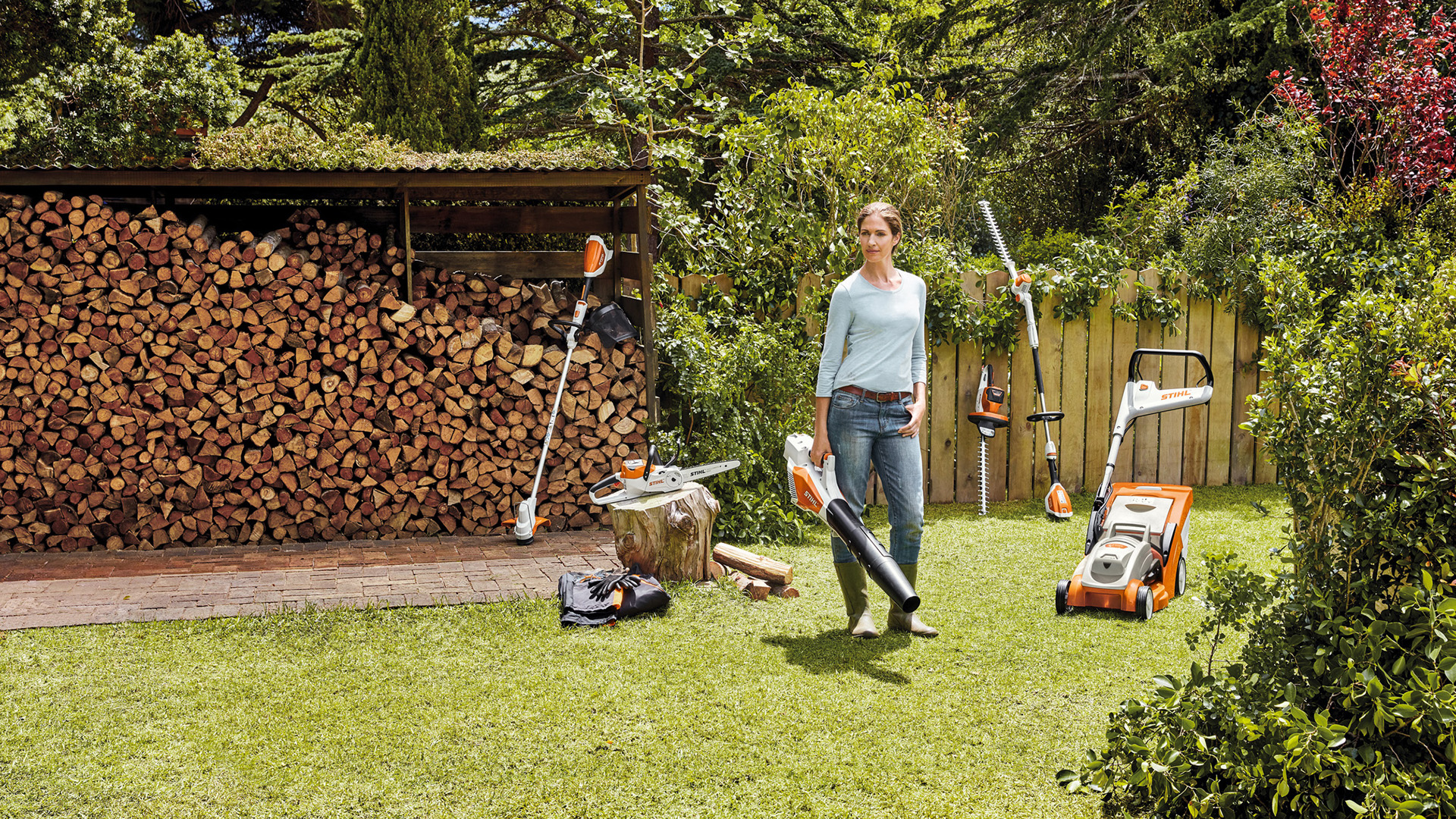 Une femme debout dans un jardin devant une sélection d’outils à batterie du système AK STIHL 