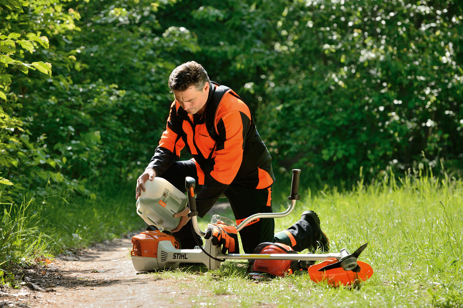 Un homme remplissant le réservoir d’une débroussailleuse STIHL à partir d’un gros bidon avec des arbustes derrière lui