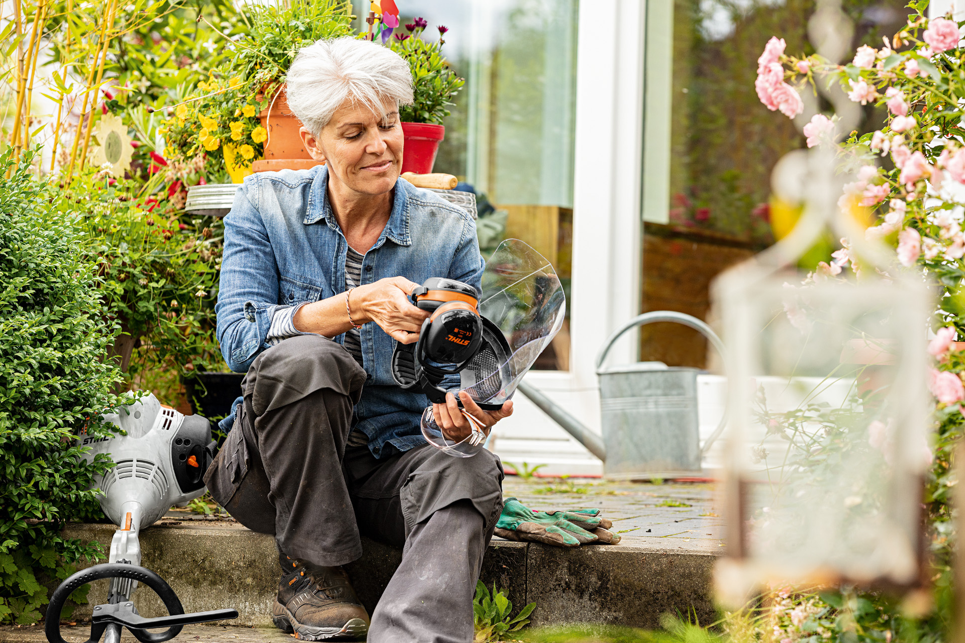 Une femme est assise entre des plantes en fleurs à côté d’une débroussailleuse STIHL FS 38 et examine l’équipement de protection