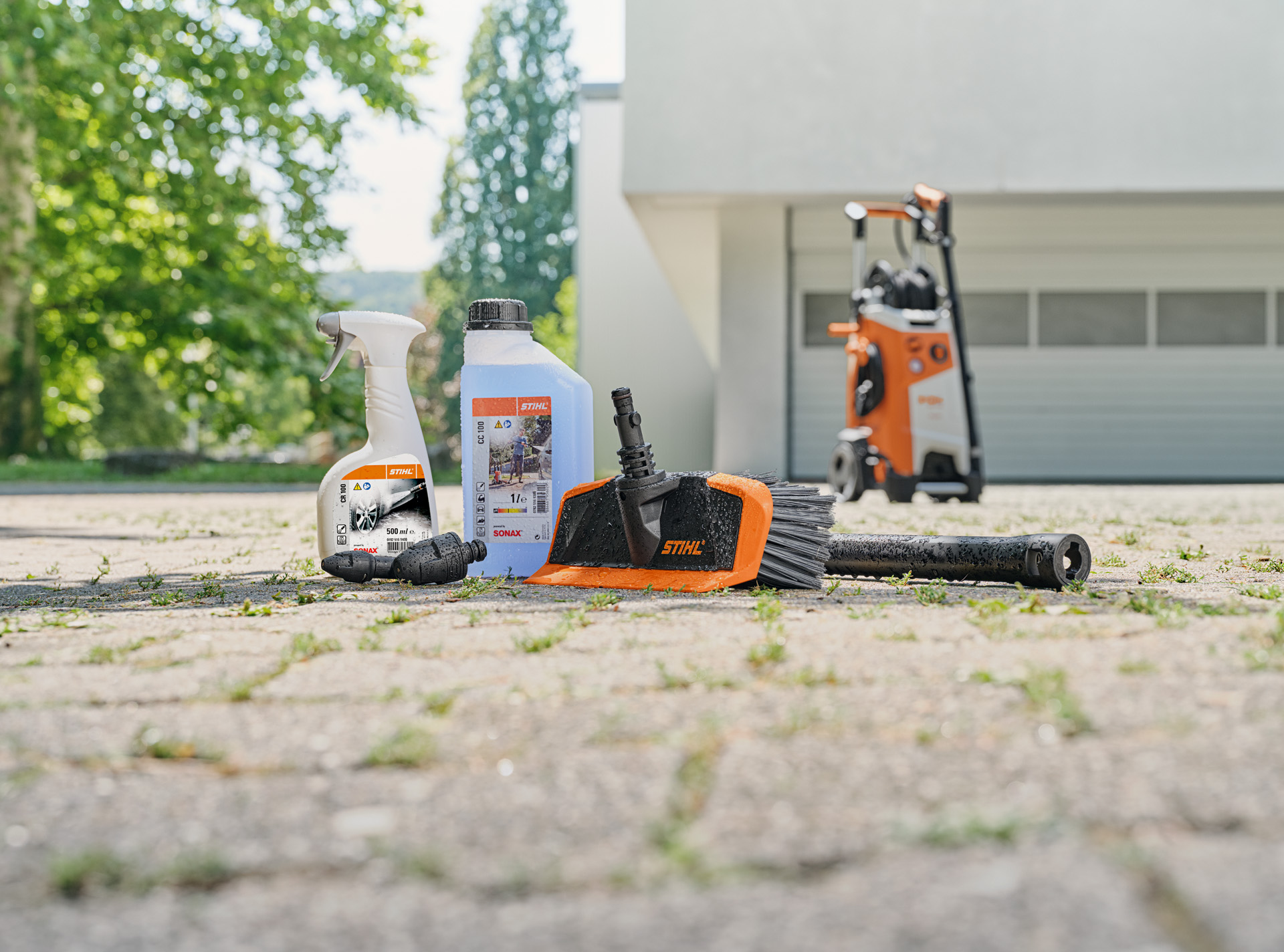  Nettoyant pour jantes STIHL, shampooing pour véhicules et brosse de lavage sur le sol devant un garage blanc. Un nettoyeur haute pression STIHL en arrière-plan