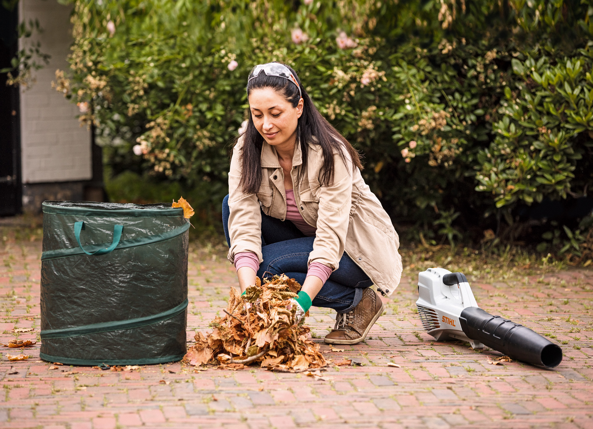 Une femme, à côté d’un souffleur de feuilles STIHL BGA 56 posé au sol, met des feuilles dans un sac à déchets végétaux