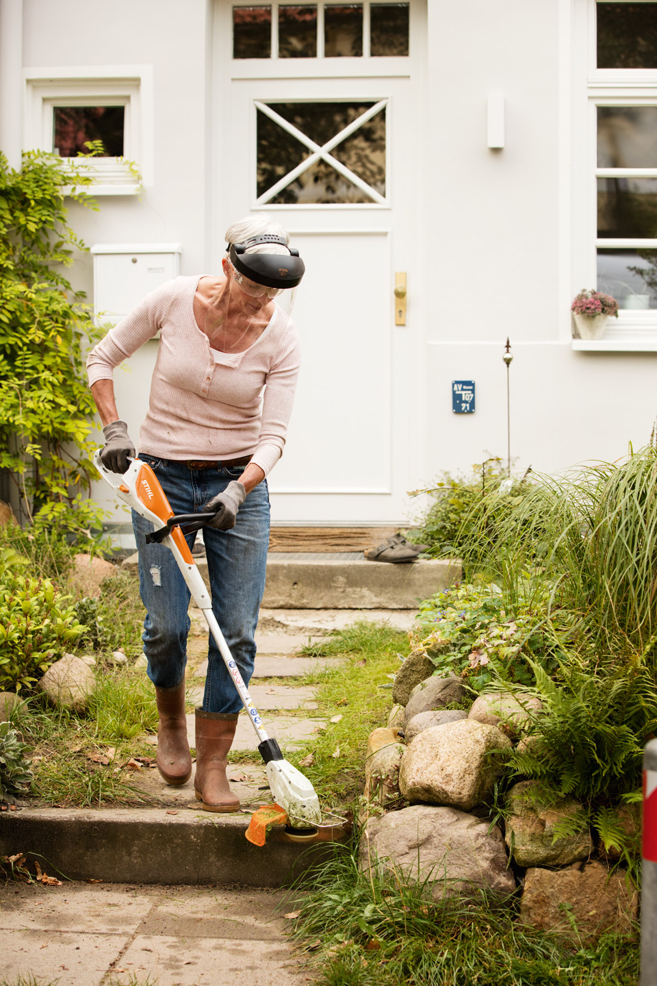 Femme avec visière de protection et gants de travail, utilisant un coupe-herbe à batterie STIHL FSA 45 devant la maison.