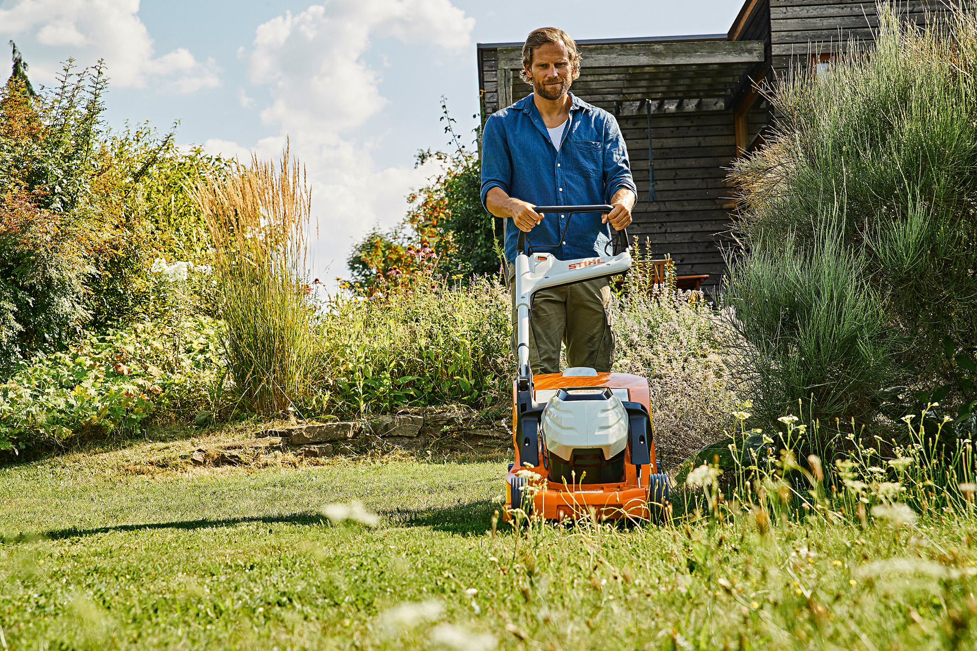 Un homme tond une pelouse à l’herbe haute avec une tondeuse sur batterie STIHL RMA 448 TC, dans un jardin