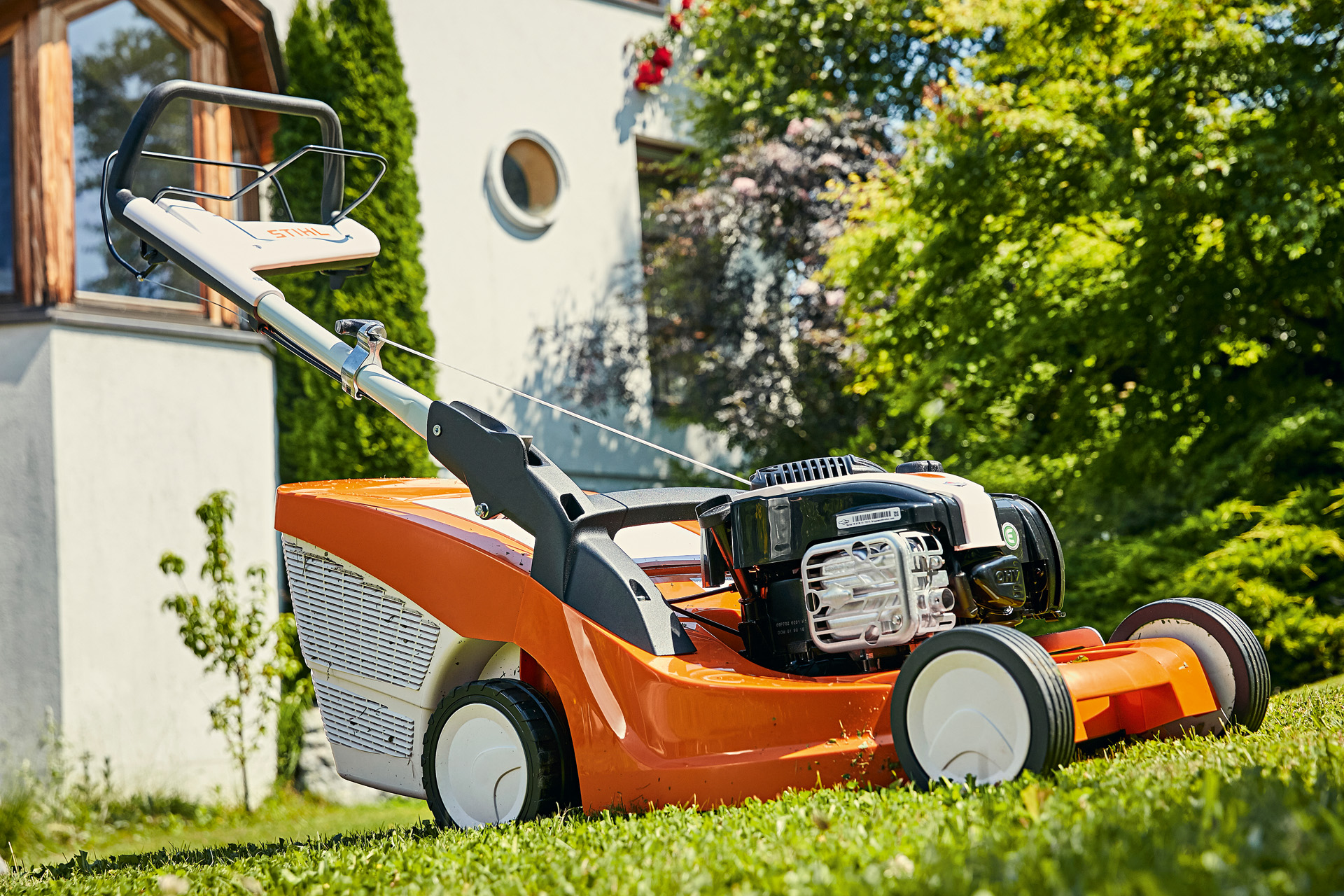 Une tondeuse à gazon thermique tractée STIHL RM 448 TC dans un jardin avec une maison en arrière plan