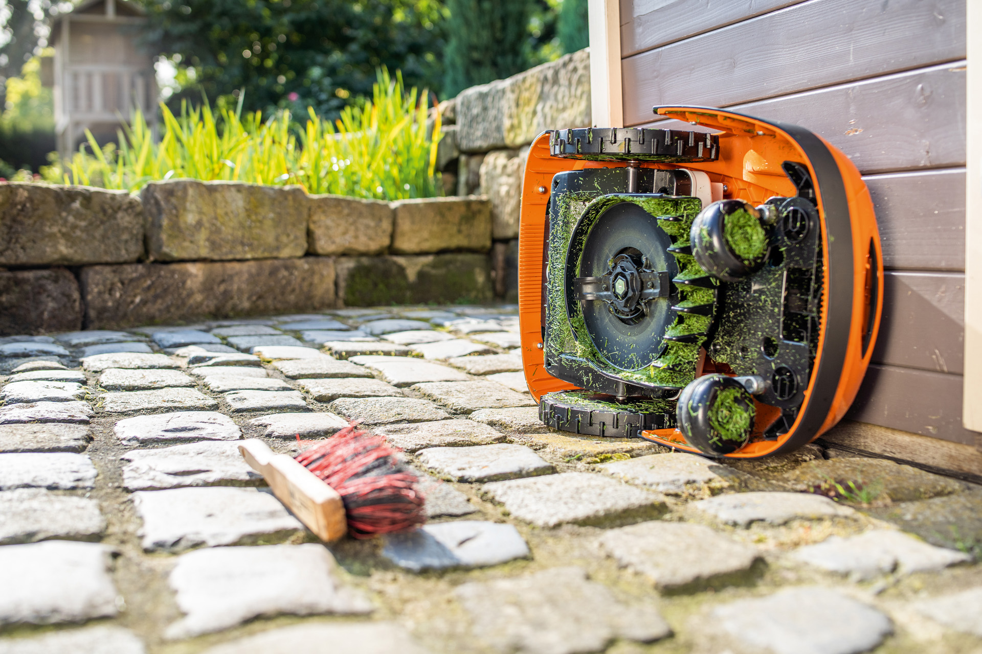 Un robot de tonte STIHL iMOW® tourné sur le côté, prêt à être nettoyé, avec une brosse posée juste à côté sur le sol