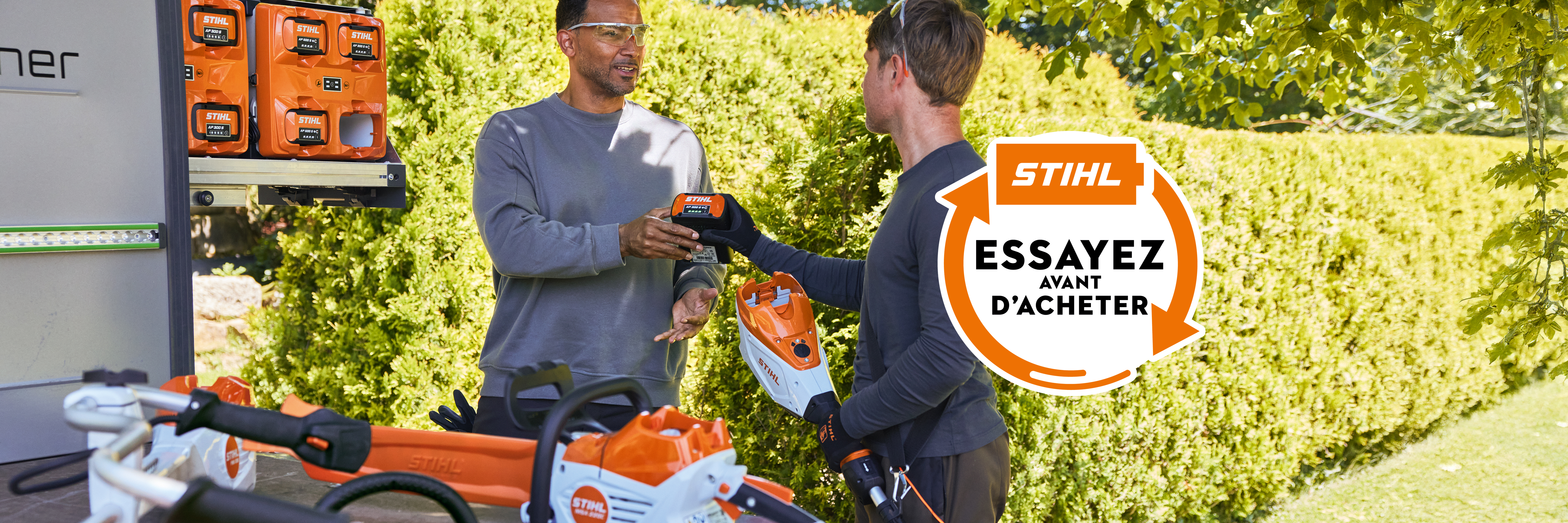 Deux hommes insérant une AP 300 S dans un taille-haies à batterie. Autocollant « Try before Buy » avec l’invitation « Test it Now – contact us for details »