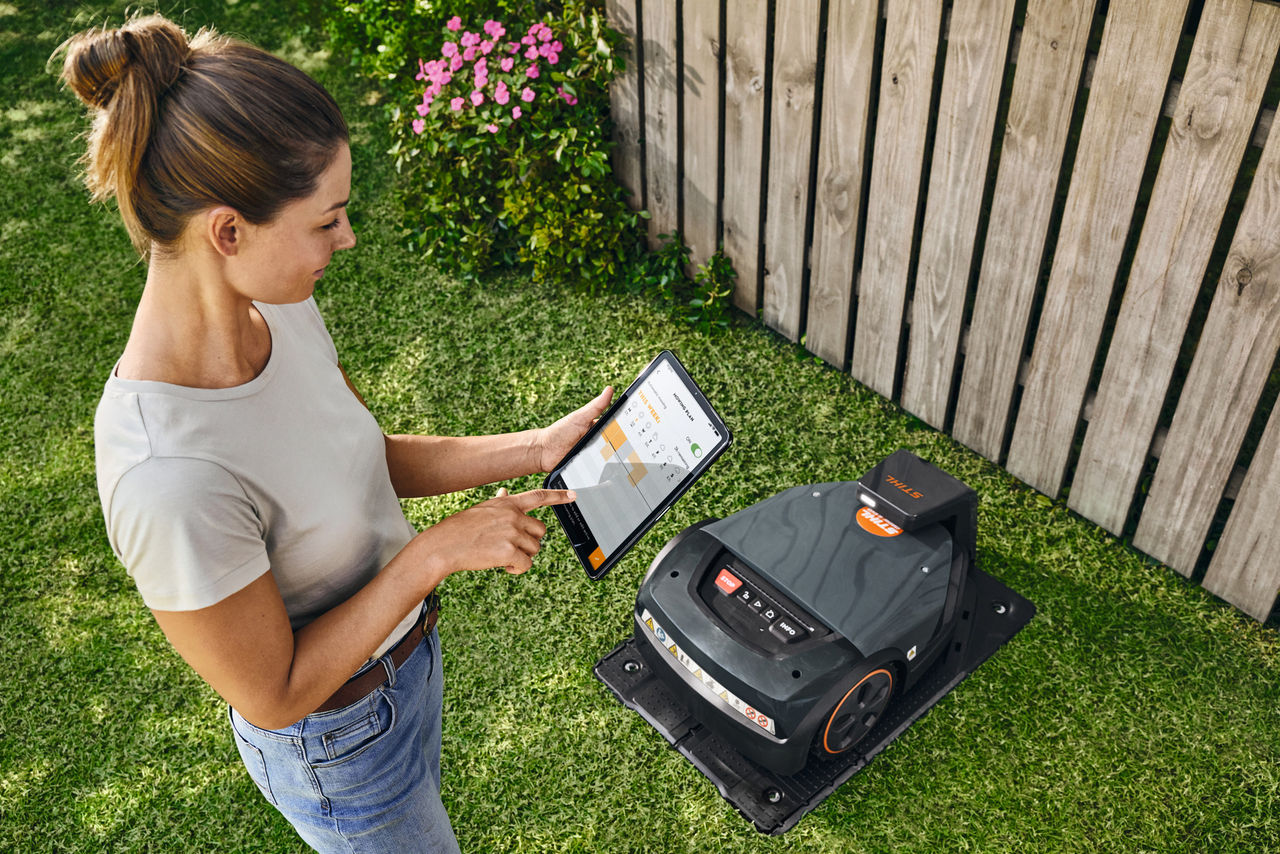 Une femme se tient devant un robot de tonte STIHL iMOW® sur la pelouse et manipule une tablette avec l’application iMOW® ouverte 