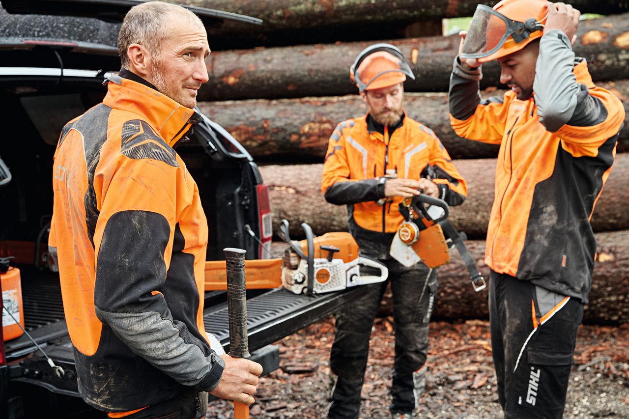 Trois hommes en équipement de protection STIHL avec des outils à batterie STIHL à côté du coffre ouvert d’un véhicule, en arrière-plan un tas de bois