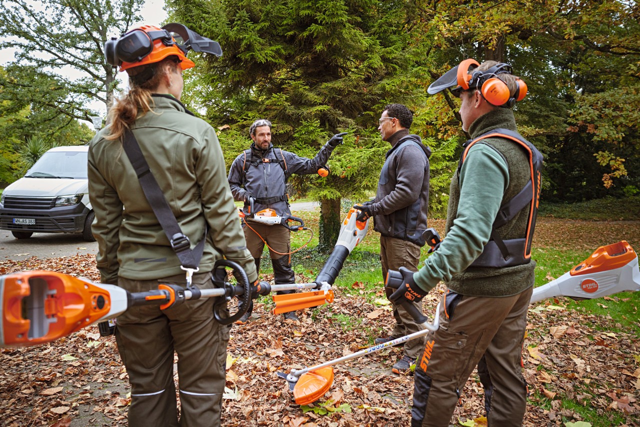 Ein Garten- und Landschaftsbauer-Team mit STIHL Profi-Akku-Geräten steht auf einer Grünfläche mit Laub im Park