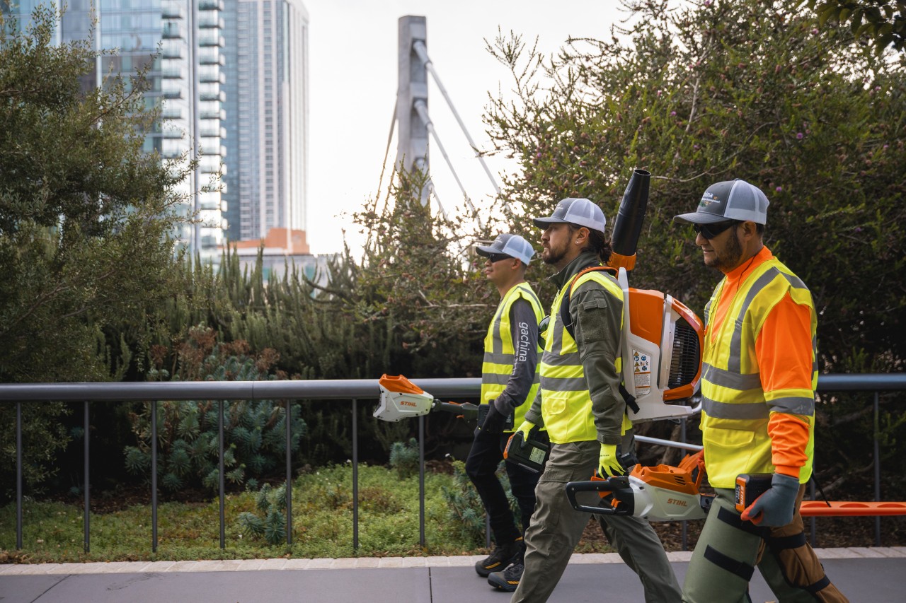 Trois travailleurs des parcs et jardins marchent en ville avec des vêtements de travail et des outils STIHL.
