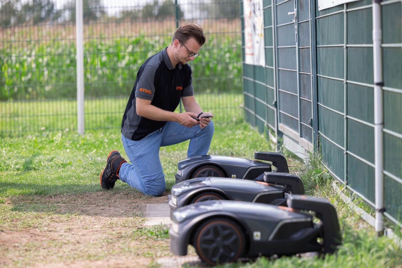 Thilo Ehrentreich, chef de produit STIHL iMOW®, est agenouillé à côté de trois robots de tonte avec un téléphone portable en main