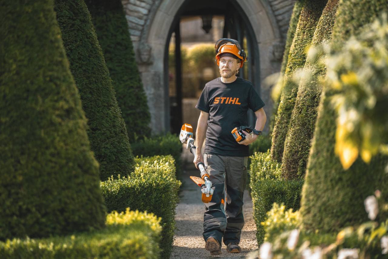 Le chef jardinier Andrew Wain traverse une allée du jardin avec un casque de protection, un T-shirt STIHL et un outil à batterie STIHL avec une batterie séparée à la main.