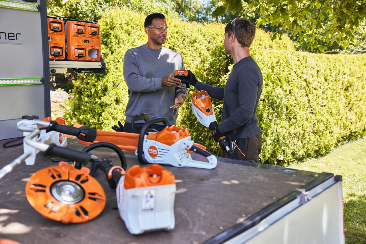 Un homme remet à son collègue une batterie STIHL, tous deux sont derrière la surface de chargement d’un véhicule, sur lequel se trouvent notamment le bottTainer et une tronçonneuse à batterie STIHL ainsi que d’autres appareils.
