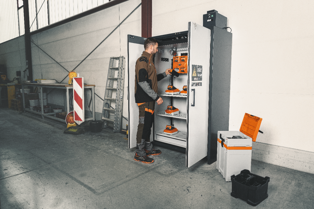 Au premier plan, une femme blonde en tenue STIHL près de l’armoire de charge de batteries dans un atelier, à l’arrière-plan, un homme aux cheveux gris chargeant un véhicule.