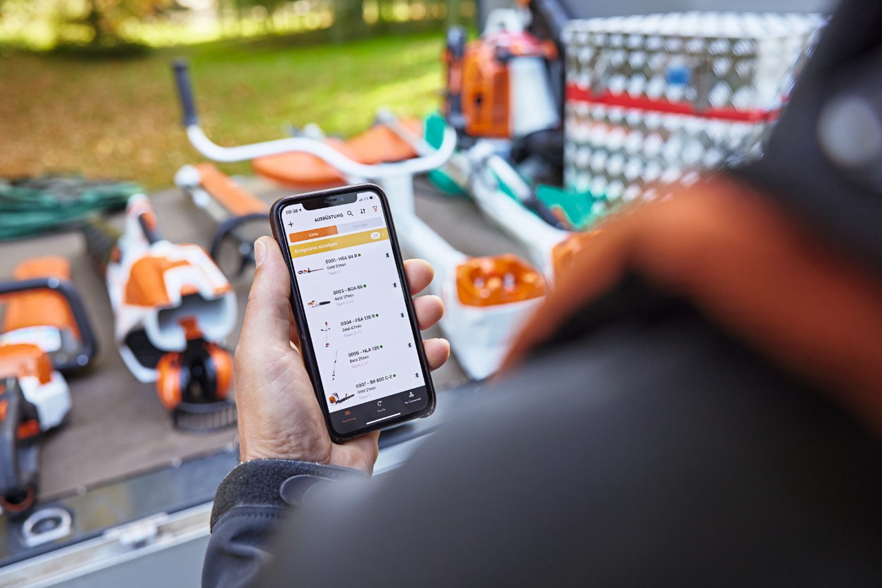 Person hält ein Smartphone in der Hand und schaut sich die Geräteliste in der STIHL connected App an, im Hintergrund sind STIHL Geräte zu sehen.