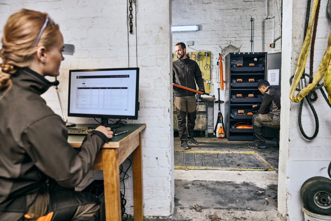 Au premier plan à gauche, une femme blonde devant un ordinateur portable dans un atelier, au fond à droite de l’image, deux hommes aux cheveux bruns devant une armoire de charge de batteries.